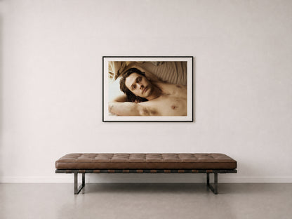 A framed 30"x 40" photography print of a reclining male portrait hangs on a wall above a brown tufted bench with black metal legs in a minimalist interior setting." 