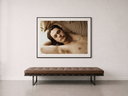 A framed art print of a shirtless man lying down, displayed on a off-white wall above a brown tufted bench.