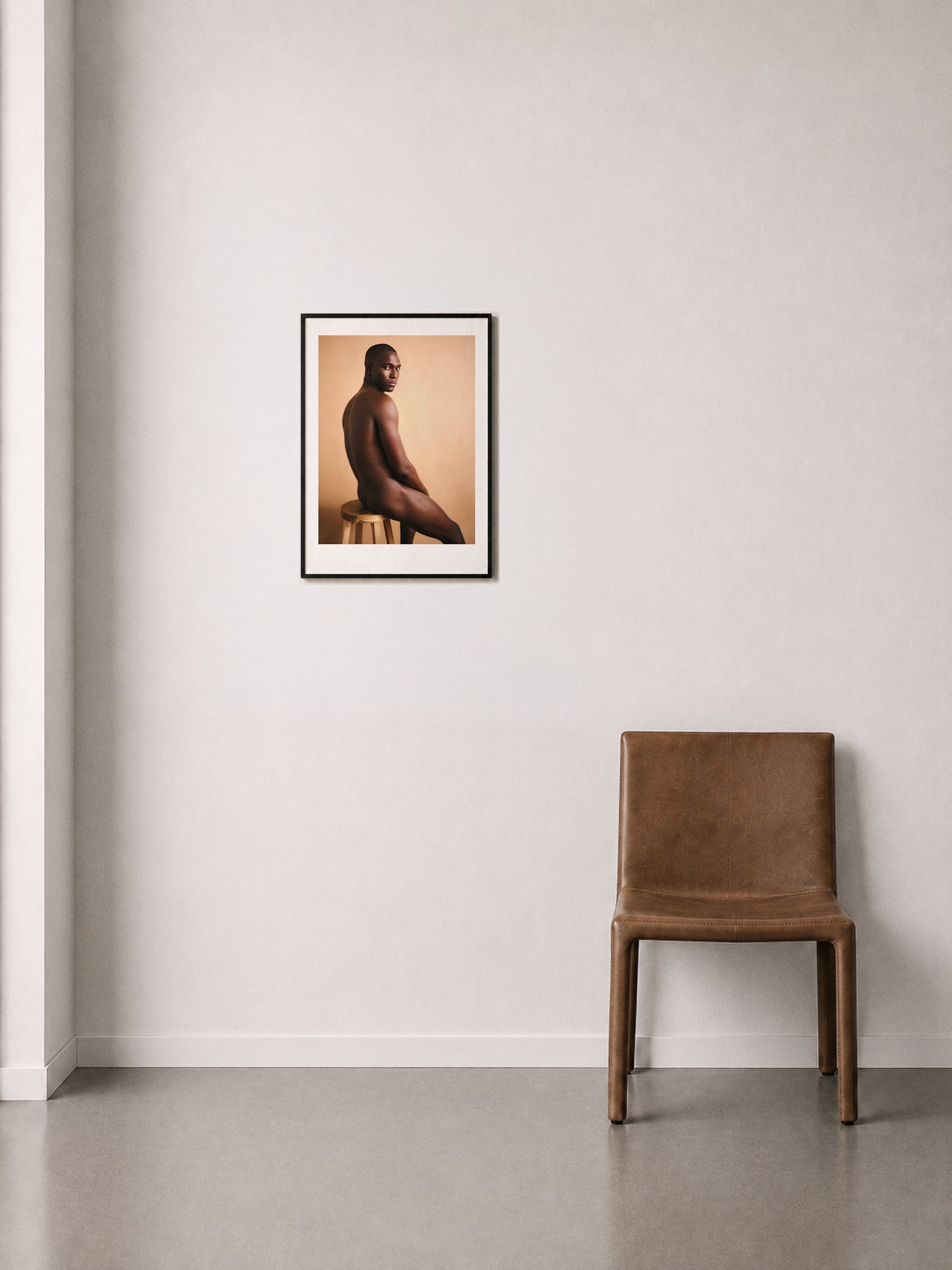 A framed art print depicting a seated nude male figure on a warm beige background, hung on a off-white wall above a brown leather chair.