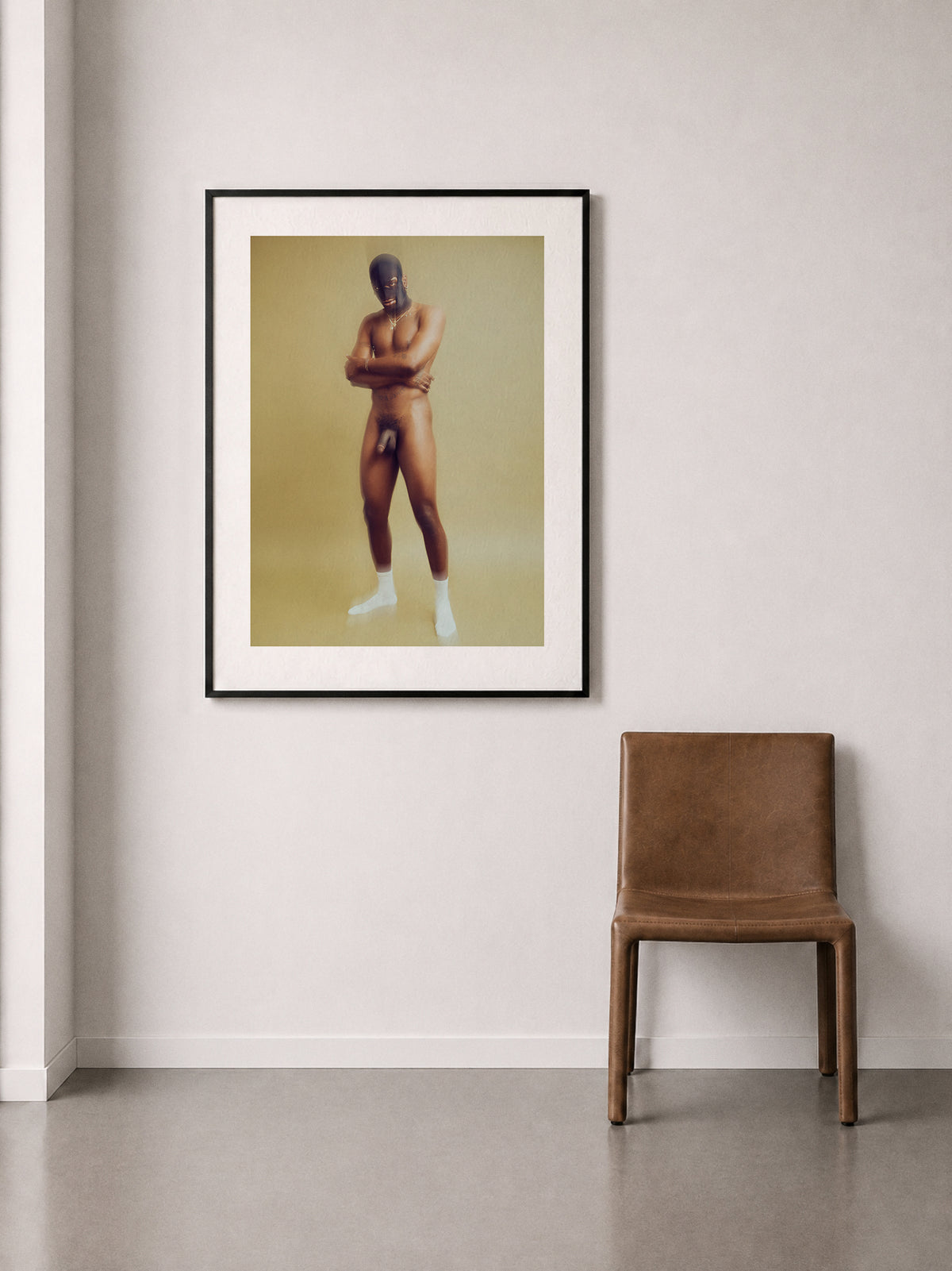A framed art print featuring a stylized portrait of a nude figure wearing a mask against a green background, displayed on a light gray wall above a brown leather chair.