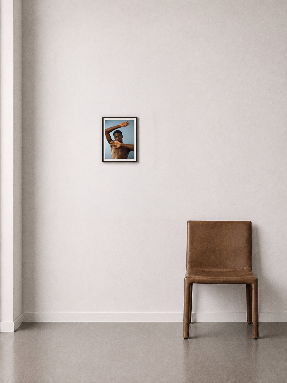A framed fine art print of a shirtless man arms in motion, against a blue background, displayed on a off-white wall above a brown leather chair.