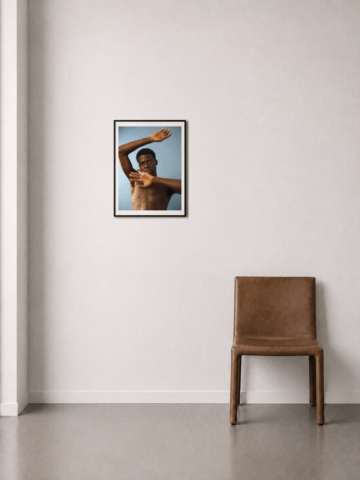 A framed fine art print of a shirtless man arms in motion, against a blue background, displayed on a off-white wall above a brown leather chair.