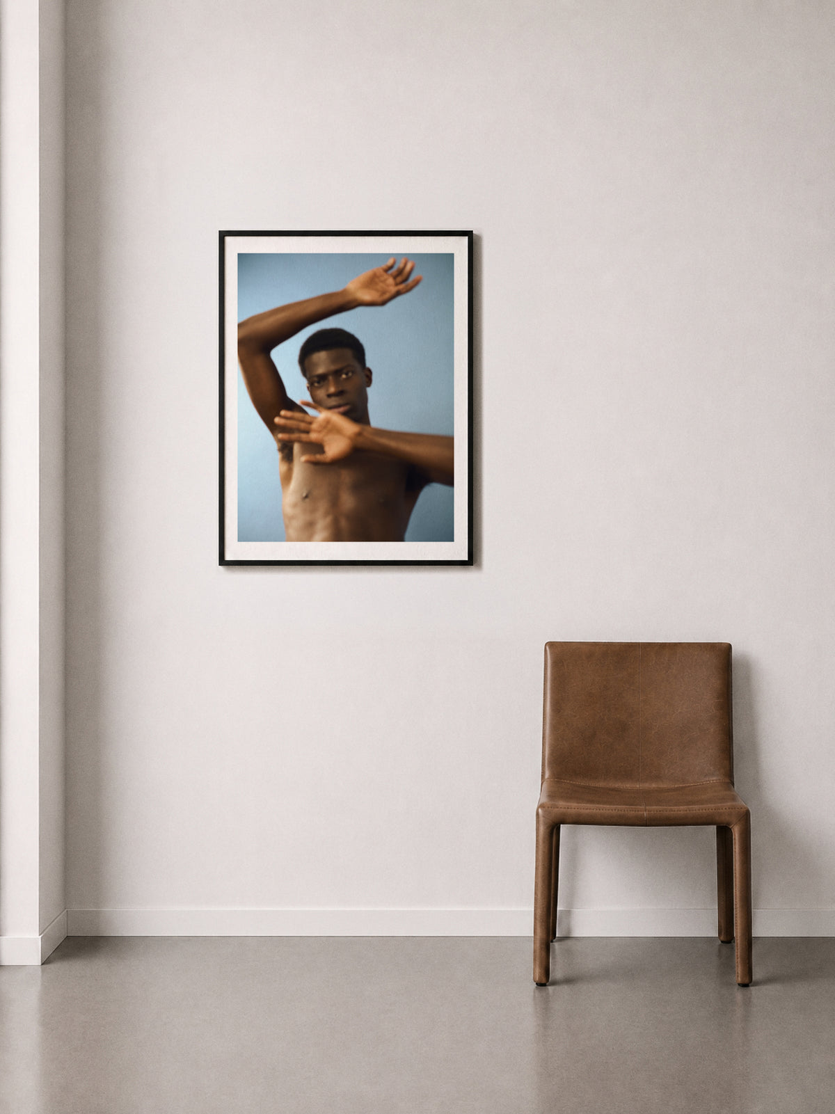 A framed fine art print of a shirtless man arms in motion, against a blue background, displayed on a off-white wall above a brown leather chair.