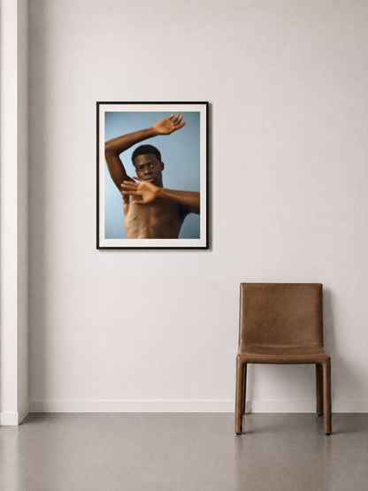 A framed fine art print of a shirtless man arms in motion, against a blue background, displayed on a off-white wall above a brown leather chair.
