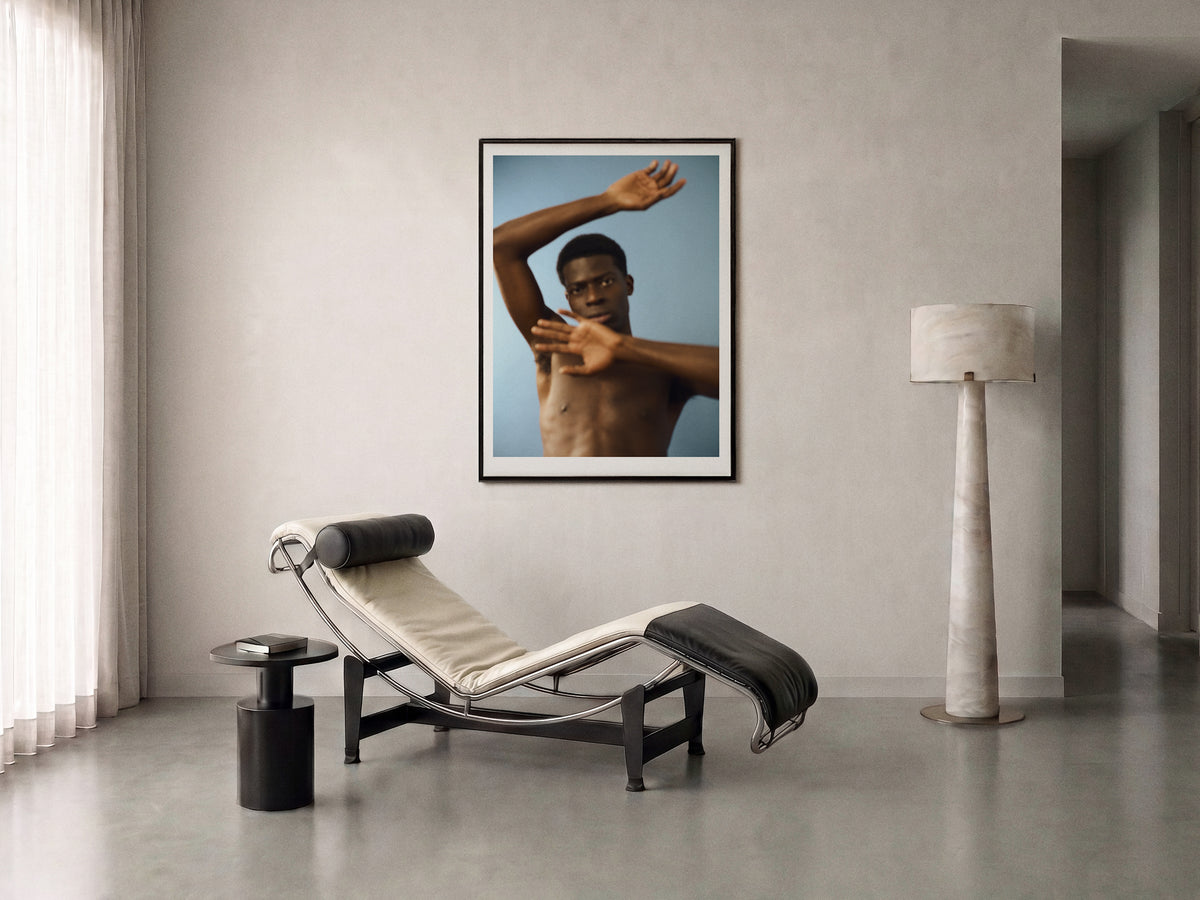 A framed fine art print of a shirtless man arms in motion, against a blue background, displayed on a off-white wall in a modern minimalist room with a chaise lounge, side table, and floor lamp.
