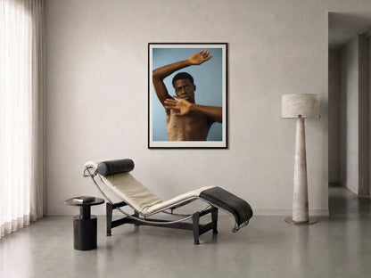 A framed fine art print of a shirtless man arms in motion, against a blue background, displayed on a off-white wall in a modern minimalist room with a chaise lounge, side table, and floor lamp.