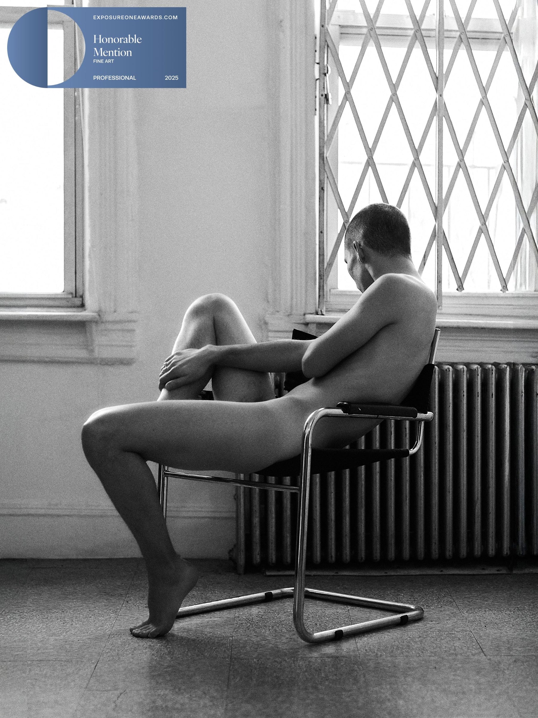 Award-winning badge of a Fine Art black and white photograph of a male nude sitting on a chair in a room with large windows.