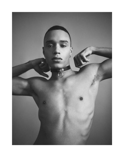 A black and white portrait of a shirtless man wearing a black choker with a silver ring, posed against a neutral background.