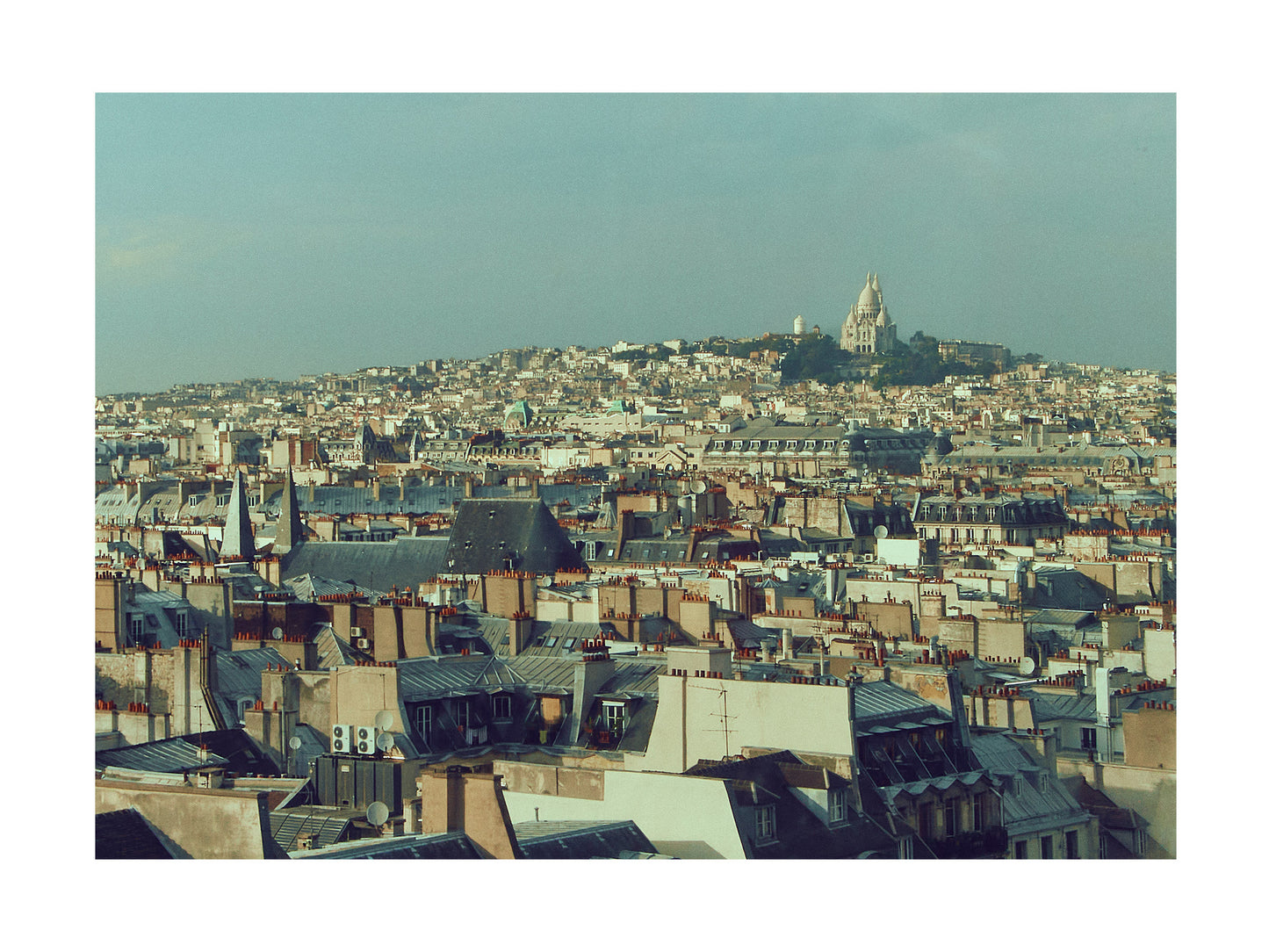Paris Cityscape with rooftops and Sacre Coeur on a hill under a clear sky.